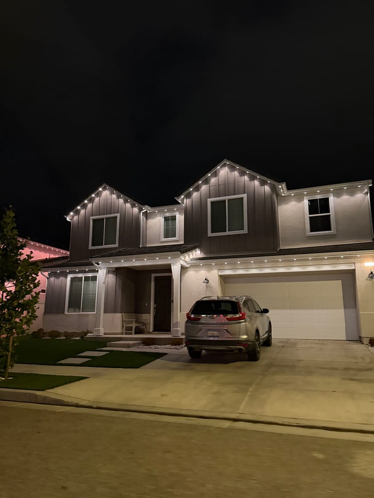 Two-story farmhouse at night with warm white permanent lights on the gables and over the garage