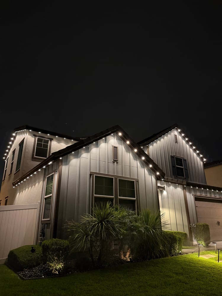 Board-and-batten home at night with bright permanent eave lights on the gables, landscape lights on the path and lit palms