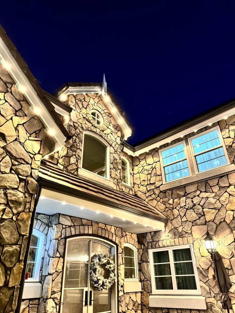 Stone home at dusk against a deep blue sky, with warm white eave lights on the gables and a wreath on the front door