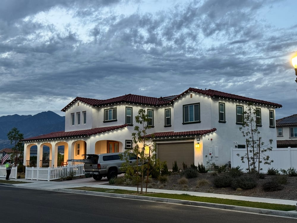 Large white stucco home with a red tile roof at dusk, warm white permanent lights on the rooflines, mountains and clouds behind and the JFX Lighting truck in the driveway