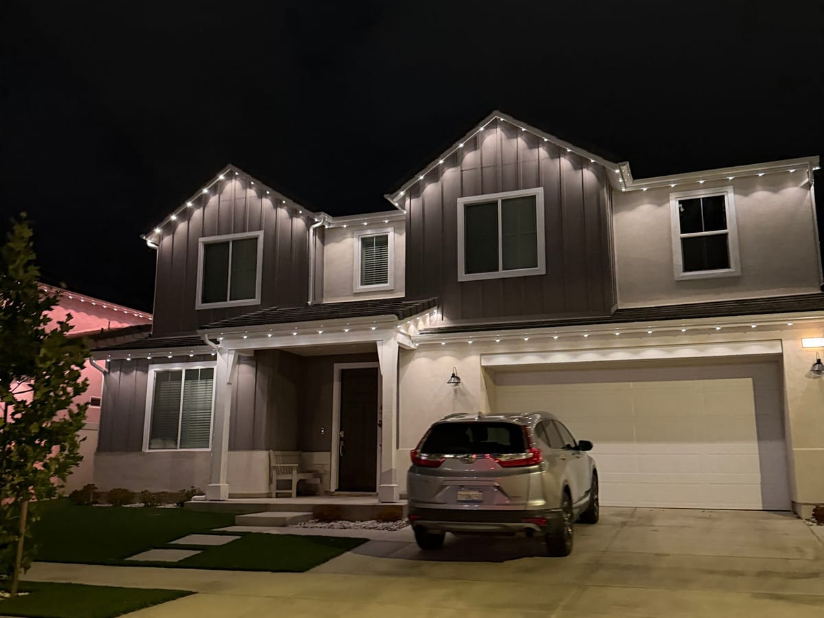 Two-story farmhouse at night with warm white permanent lights on the gables and over the garage