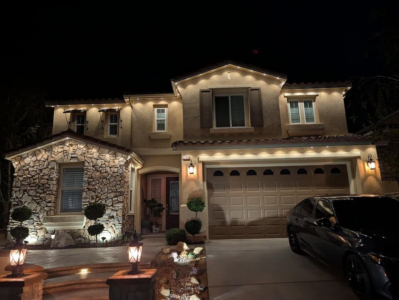 Two-story stone and stucco home at night with warm white roofline lights, lit shrubs and entry lanterns
