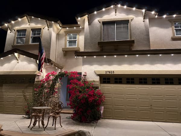 Two-story stucco home with warm white lights along the rooflines, bougainvillea and a bistro set by the front door