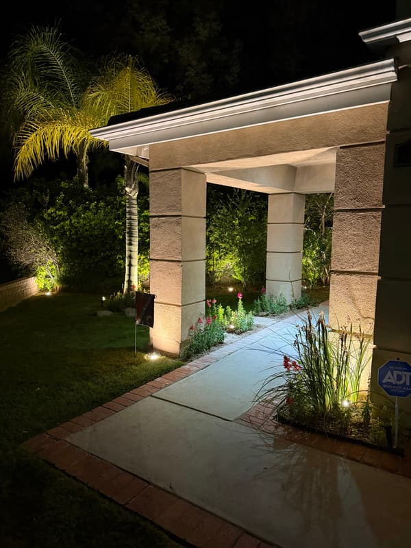 Covered entry walkway at night with uplit columns, palms and landscape lights along the planting beds