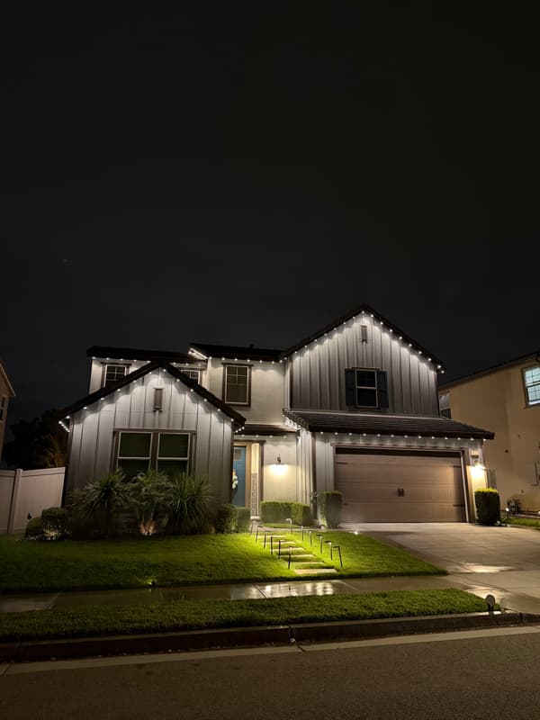 Farmhouse-style home at night with warm white roofline lights and lit landscaping