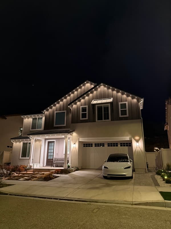 Two-story home at night with warm white lights on the gables and porch roof, a white car in the driveway