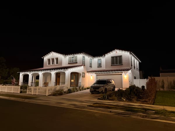 White two-story home with an arched porch and picket fence at night, warm white lights along the rooflines