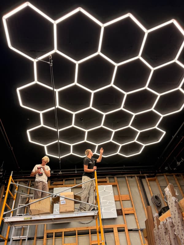 Two installers on scaffolding under a large hexagon LED ceiling in a room still under construction