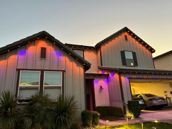 Board-and-batten home at dusk with orange and purple permanent eave lights for Halloween